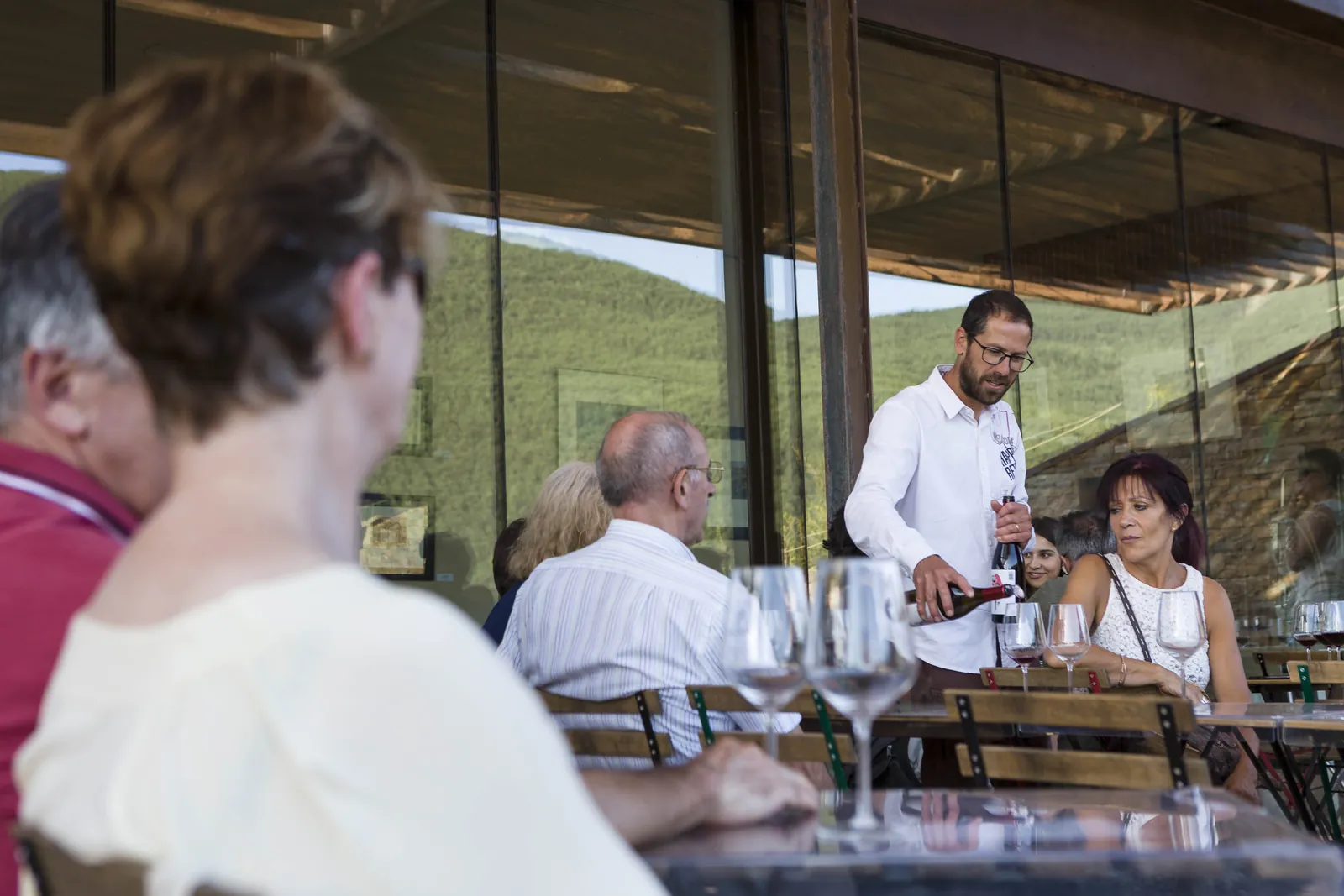 El enólogo sirve vino a un grupo sentado en la terraza de la bodega, con las cristaleras y el monte detrás, en Barbenuta